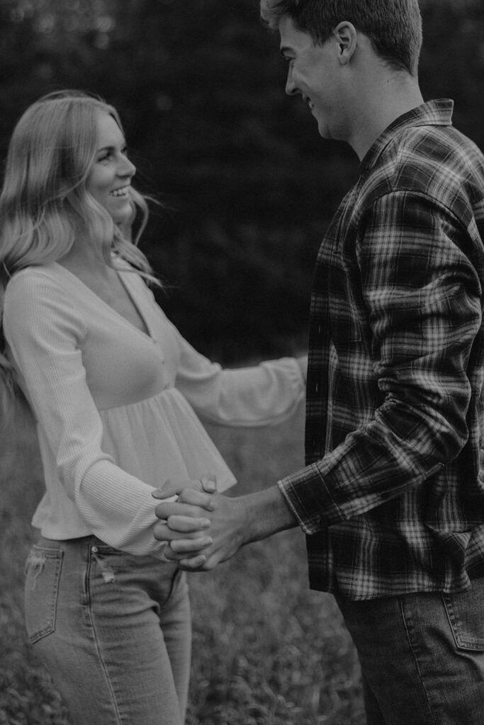 Tess and Cole holding hands and laughing together in a grassy field in Sioux Falls, surrounded by vibrant fall foliage for their park engagement photos