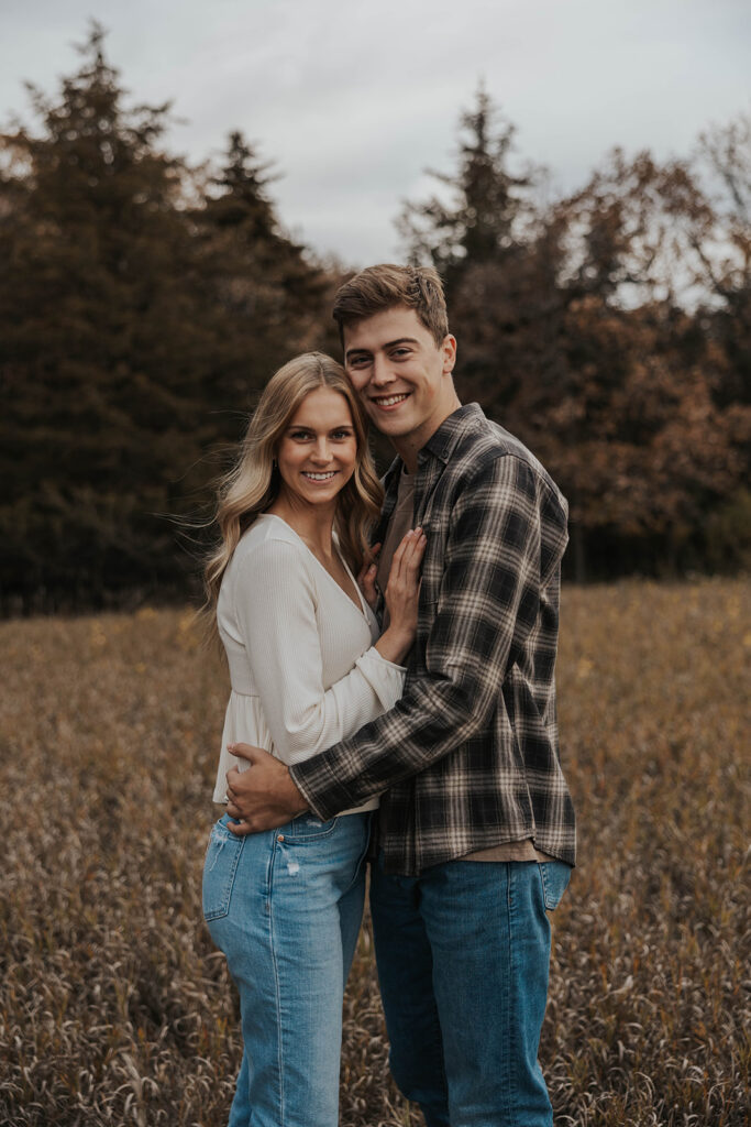Tess and Cole holding hands and laughing together in a grassy field in Sioux Falls, surrounded by vibrant fall foliage for their park engagement photos