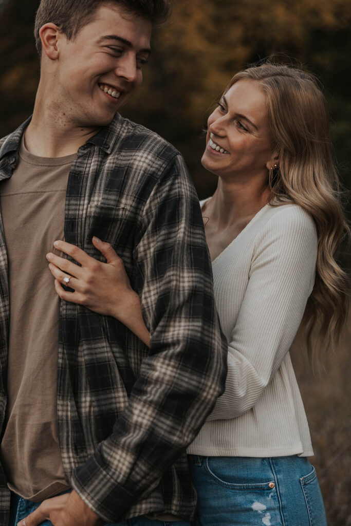 Tess and Cole hugging in a grassy field in Sioux Falls, surrounded by vibrant fall foliage for their park engagement photos