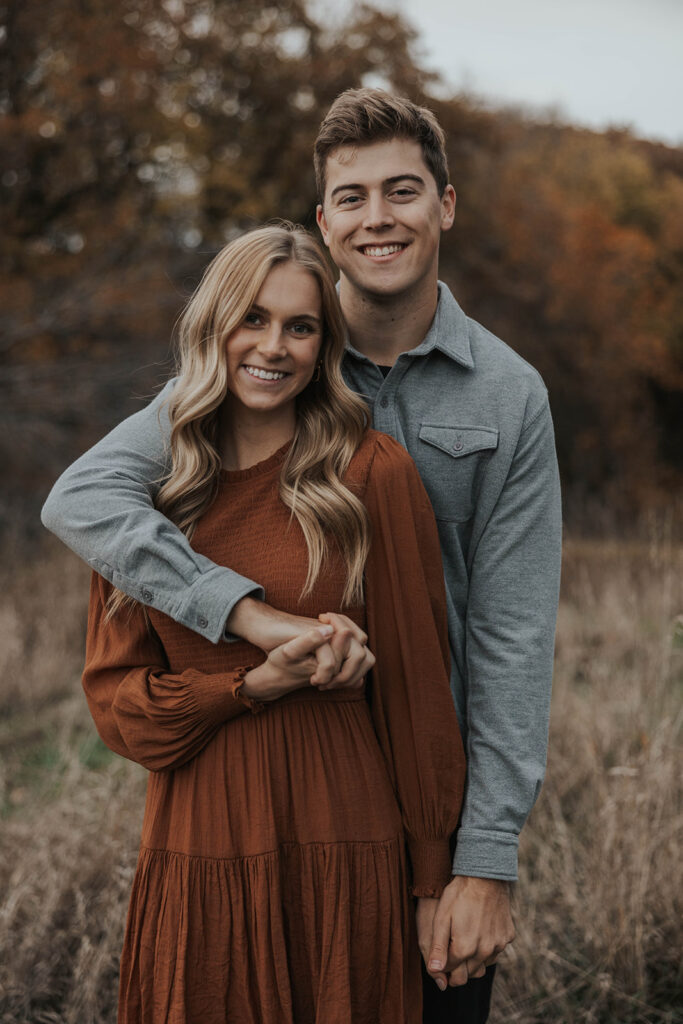 Tess and Cole holding hands and laughing together in a grassy field in Sioux Falls, surrounded by vibrant fall foliage