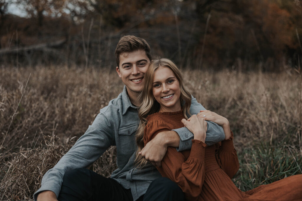 The couple sitting cozily on in a field, sharing a warm embrace and smiling at each other for their park engagement photos
