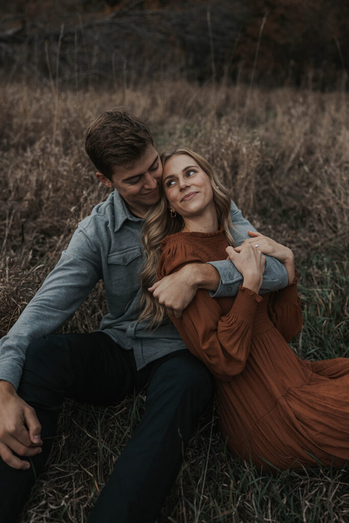 The couple sitting cozily on in a field, sharing a warm embrace and smiling at each other for their park engagement photos