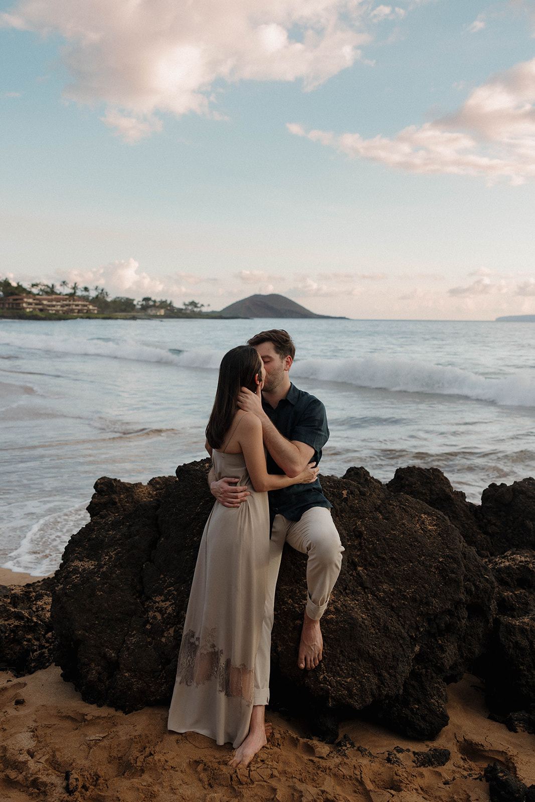 a man sitting on a rock and a woman standing as she kisses him in Maui 