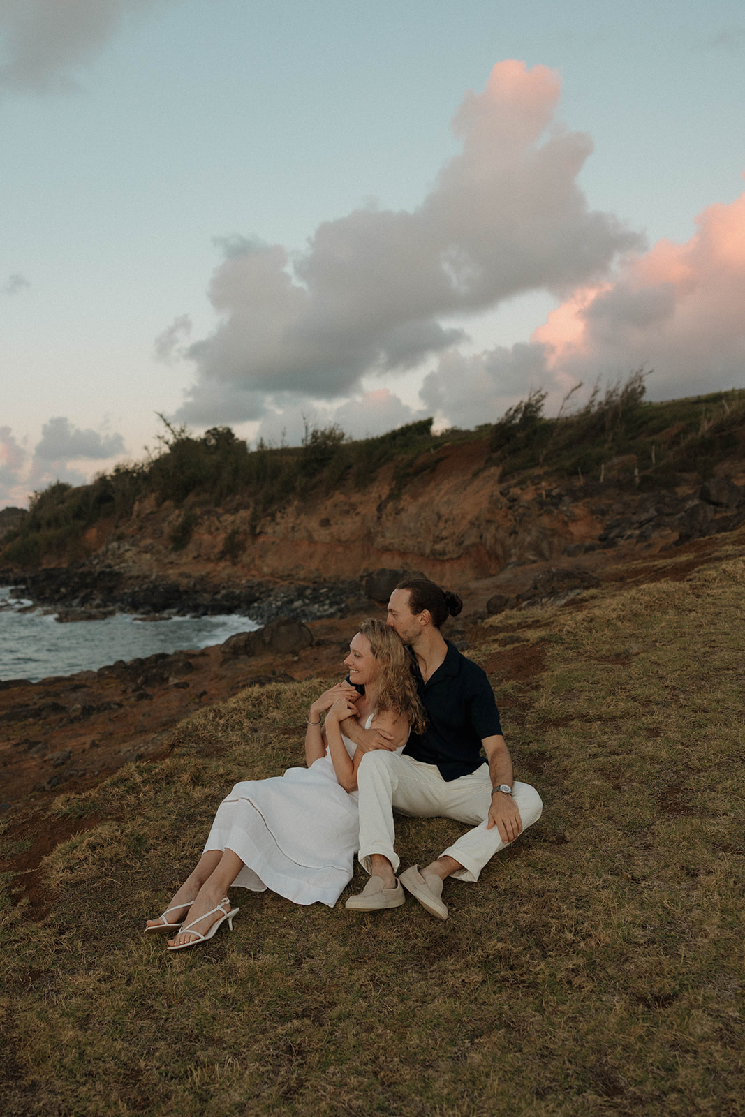 a couple sitting in the grass, looking out to the ocean, captured by a Maui proposal photographer
