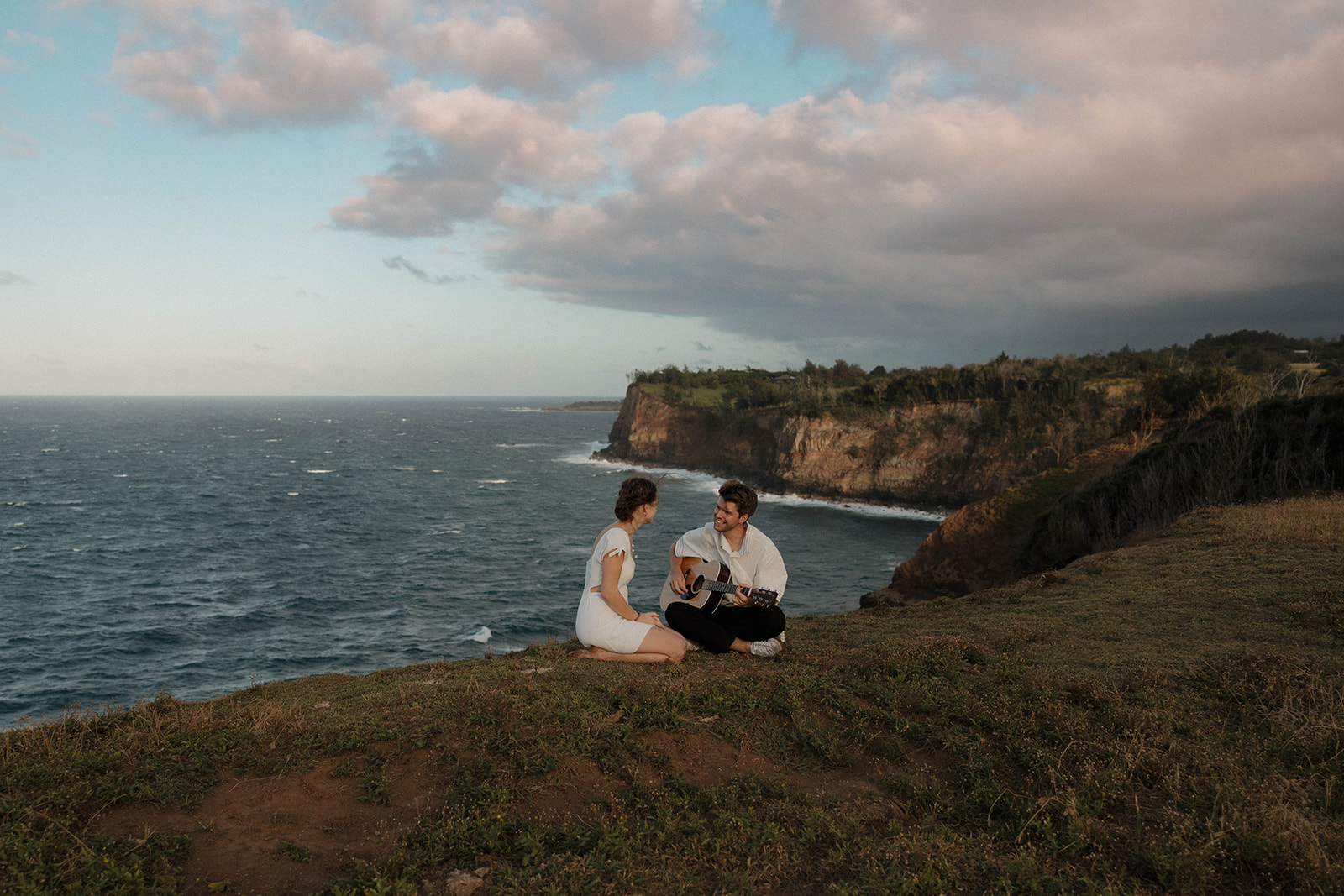 a man playing guitar as a woman listens as they sit near a cliff in Hawaii 