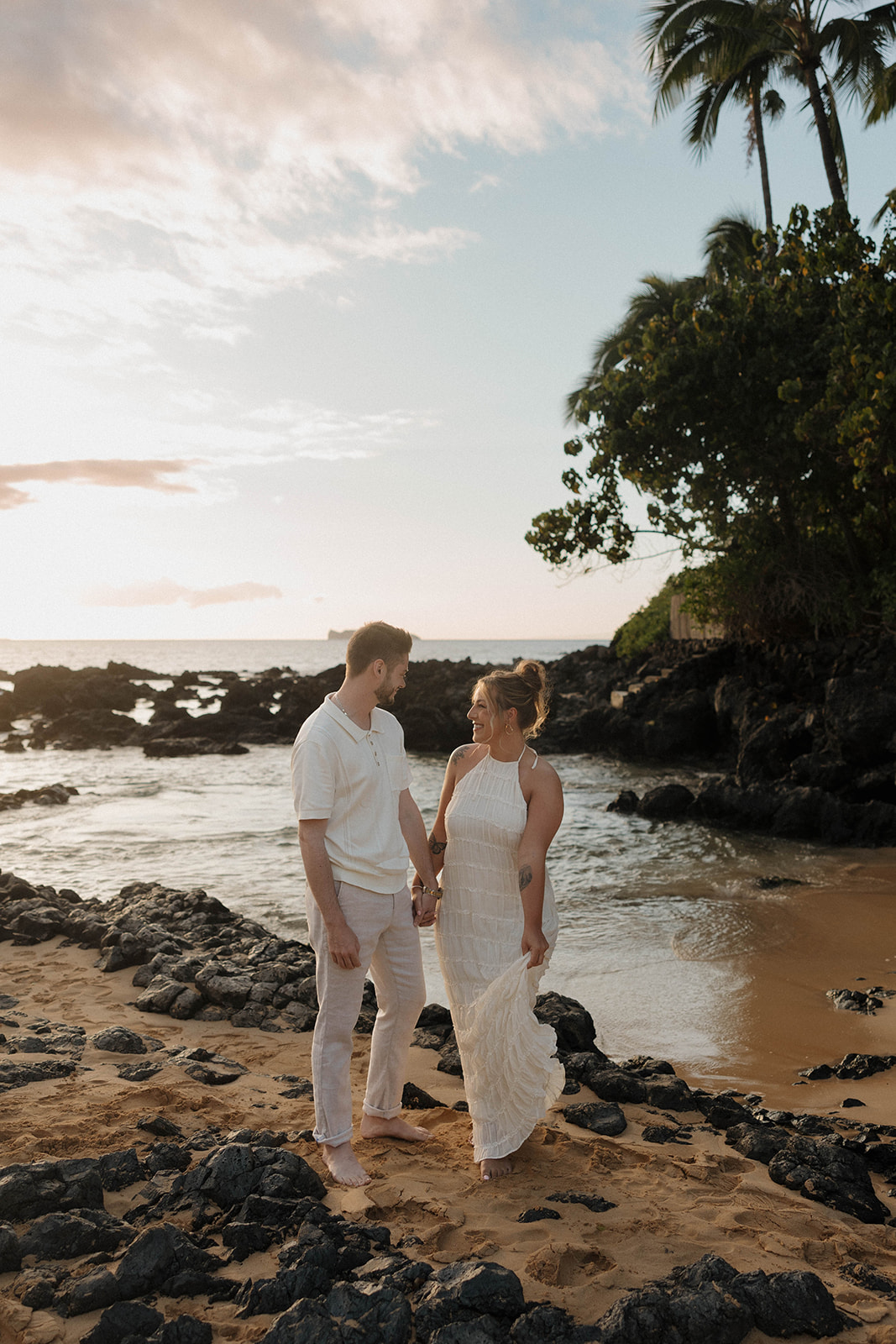 a couple smiling together on the beach during golden hour as they wear beautifully coordinated engagement photo outfits 