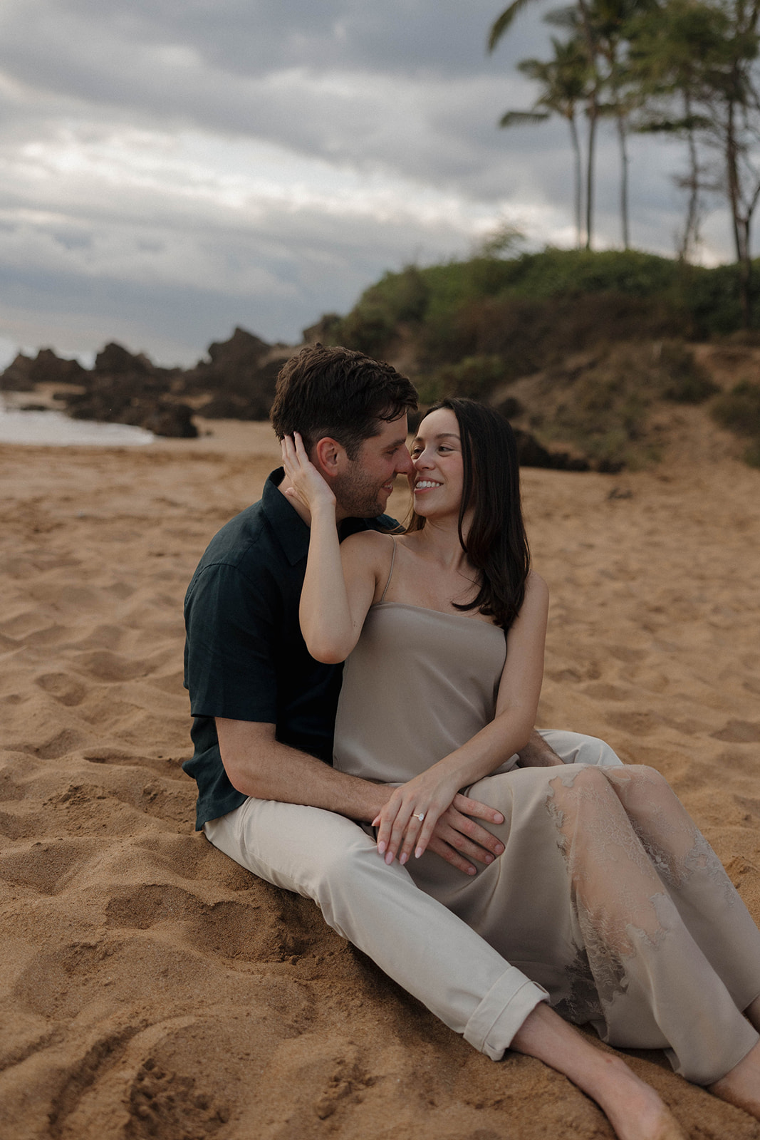 a couple sitting in the sand on the beach showcasing what to wear for engagement photos