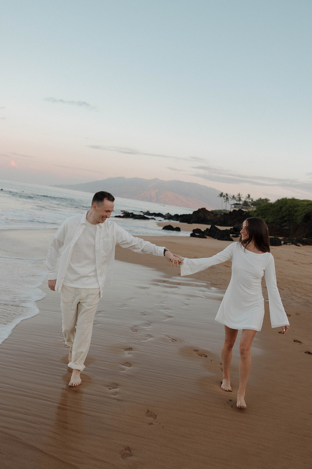 a couple wearing white outfits as they walk on the beach as they feature what to wear for engagement photo outfits
