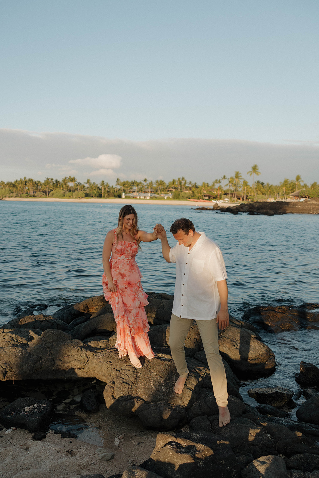 a man helping a woman down from some rocks by the ocean during their engagement session