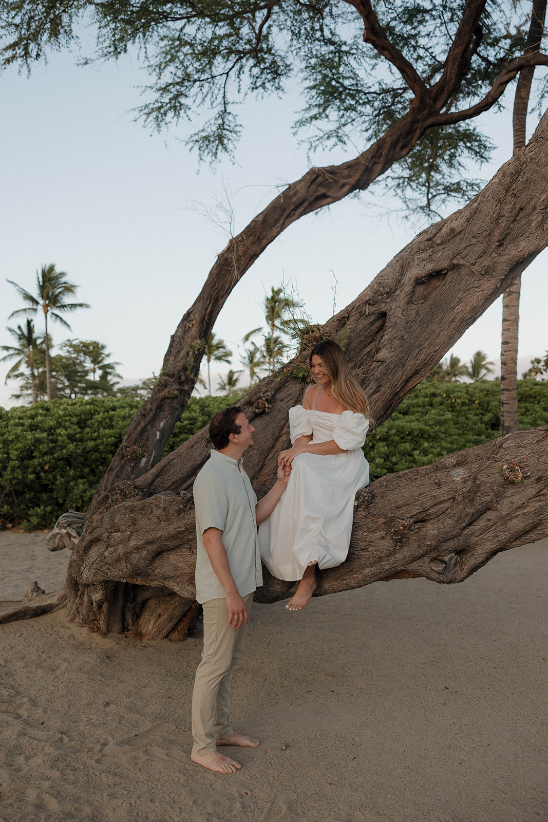 a woman in a white dress sitting on a tree and a man standing beside her showing ideas on engagement photo outfits