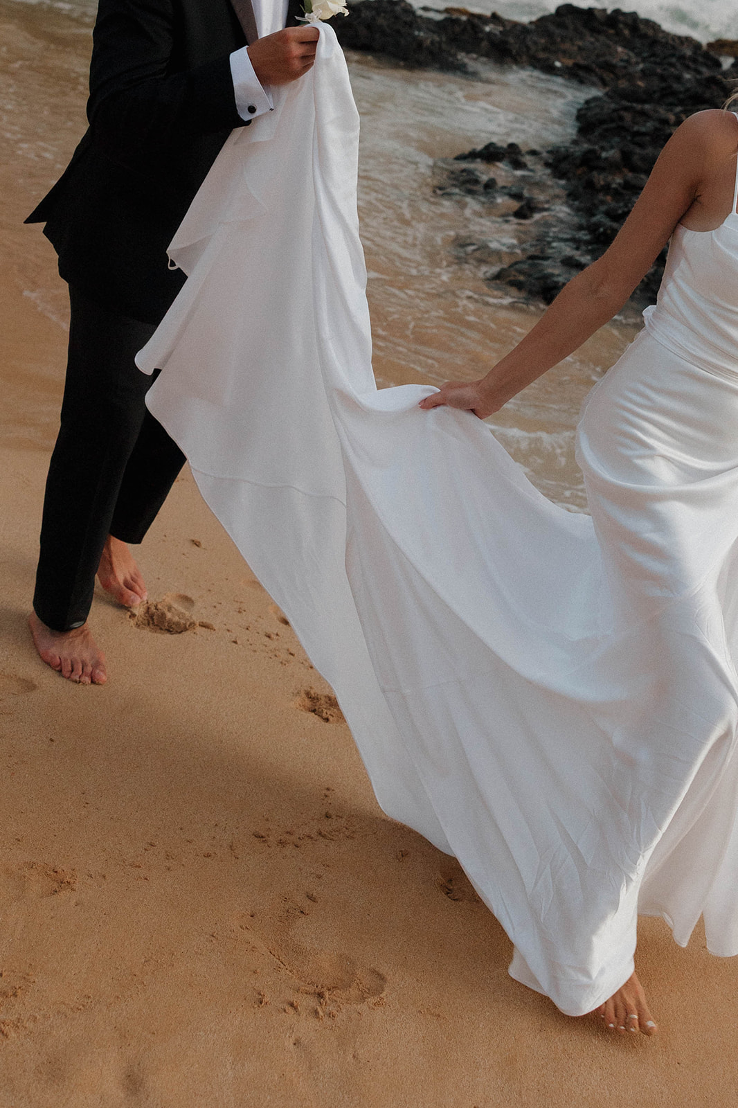 a groom holding the back of a bride's dress as they walk on the beach in Maui 