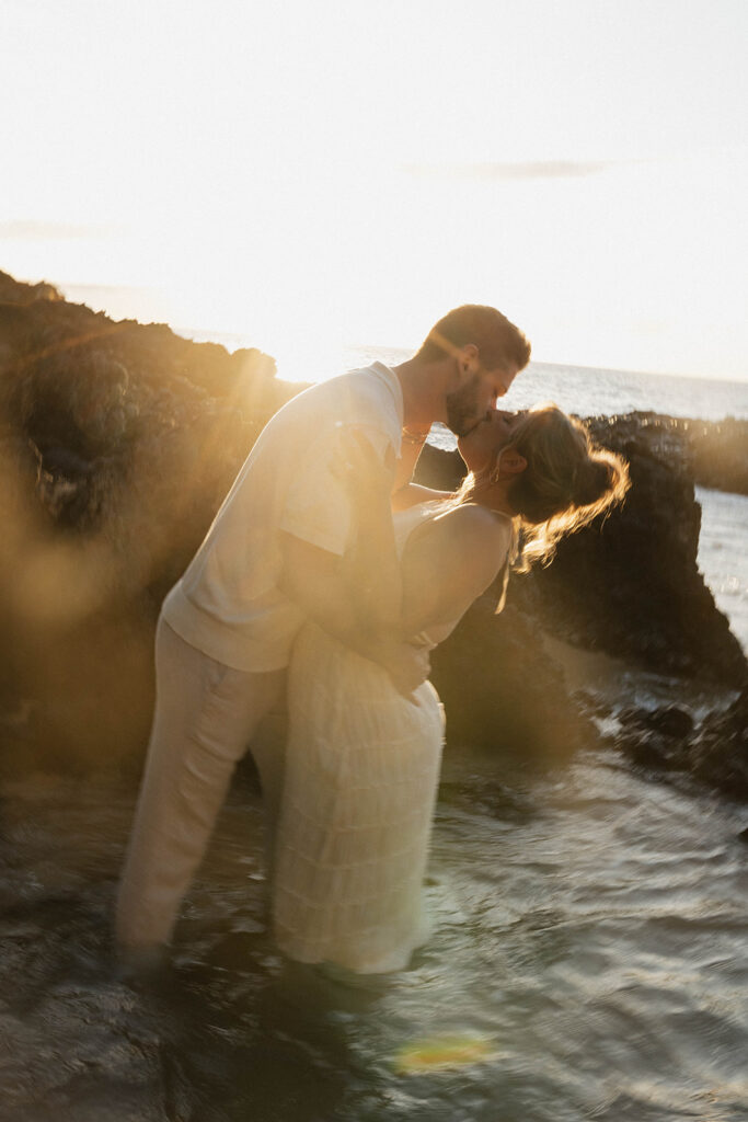 A gorgeous photo of a couple standing in the waves with the sunset and black rocks behind them during their engagement shoot.