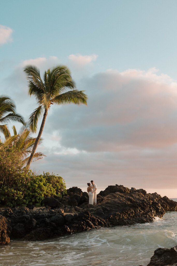 A wide photo of a couple standing on black rocks on the coast in Maui during their beach engagement shoot.