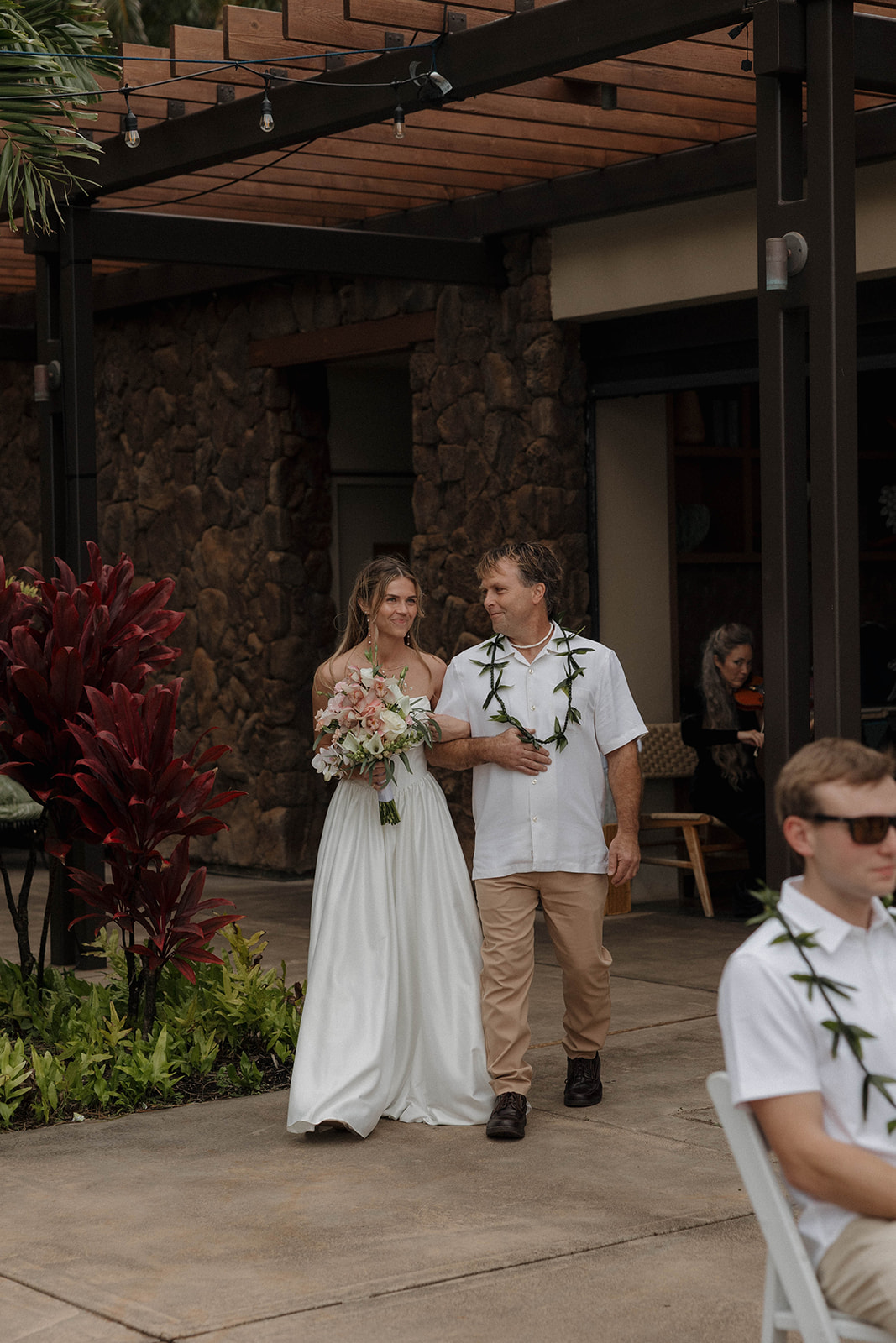 Bride walking down the aisle with her father under a wood pergola surrounded by lush greenery and guests seated outdoors.