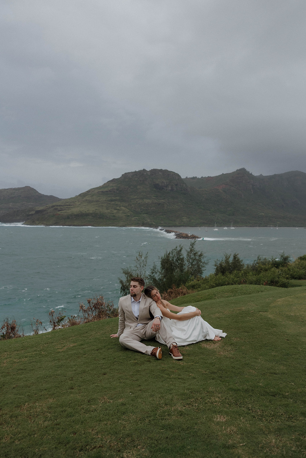 Bride and groom sitting together on a grassy bluff with waves crashing below them.