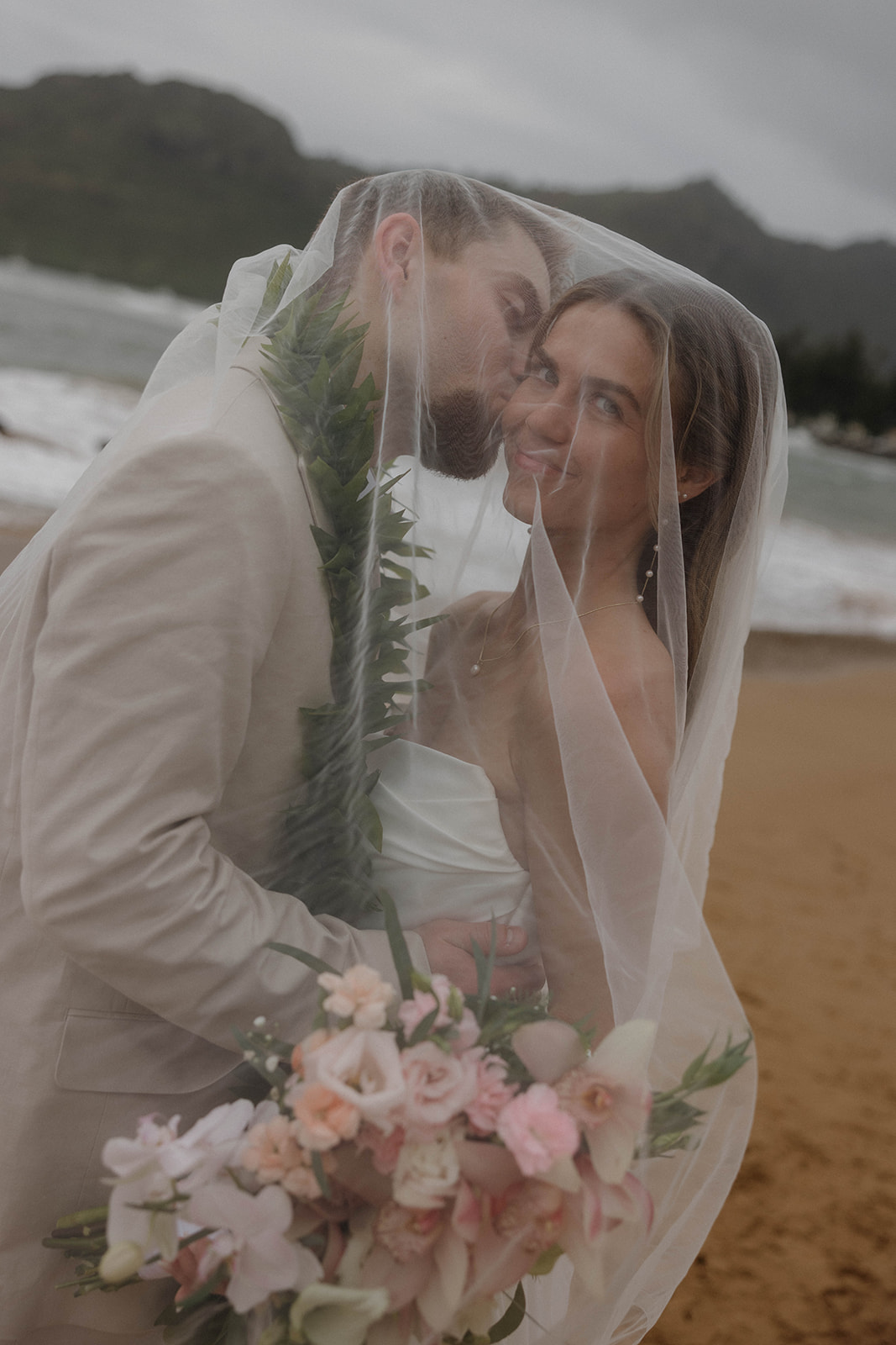 Close-up veil portrait of the bride smiling as the groom kisses her cheek on the beach.