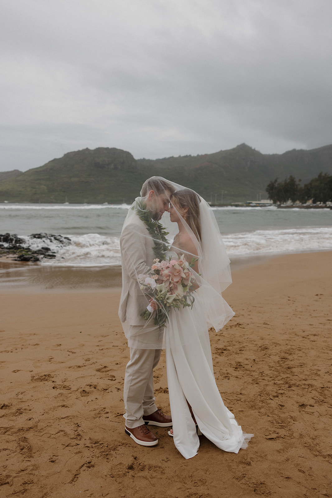 Outdoor beach portraits of the bride and groom embracing under her veil with misty mountains behind them during a destination wedding in hawaii.