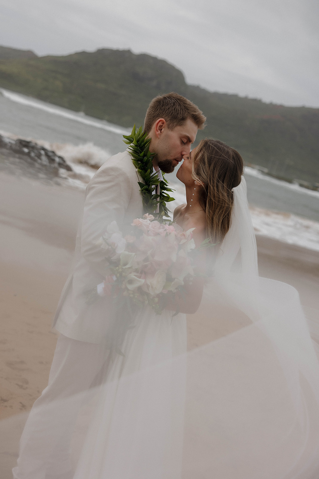 Outdoor beach portraits of the bride and groom embracing with the brides veil flowing in the foreground with misty mountains behind them during a destination wedding in hawaii.