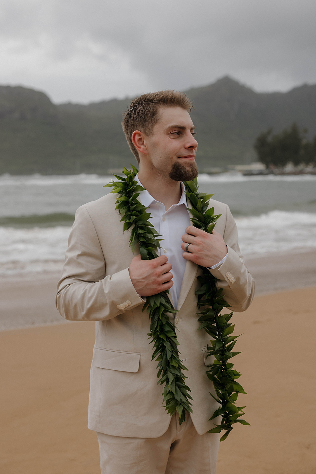 Groom wearing a beige suit and green lei posing along the shoreline during a destination wedding in hawaii.