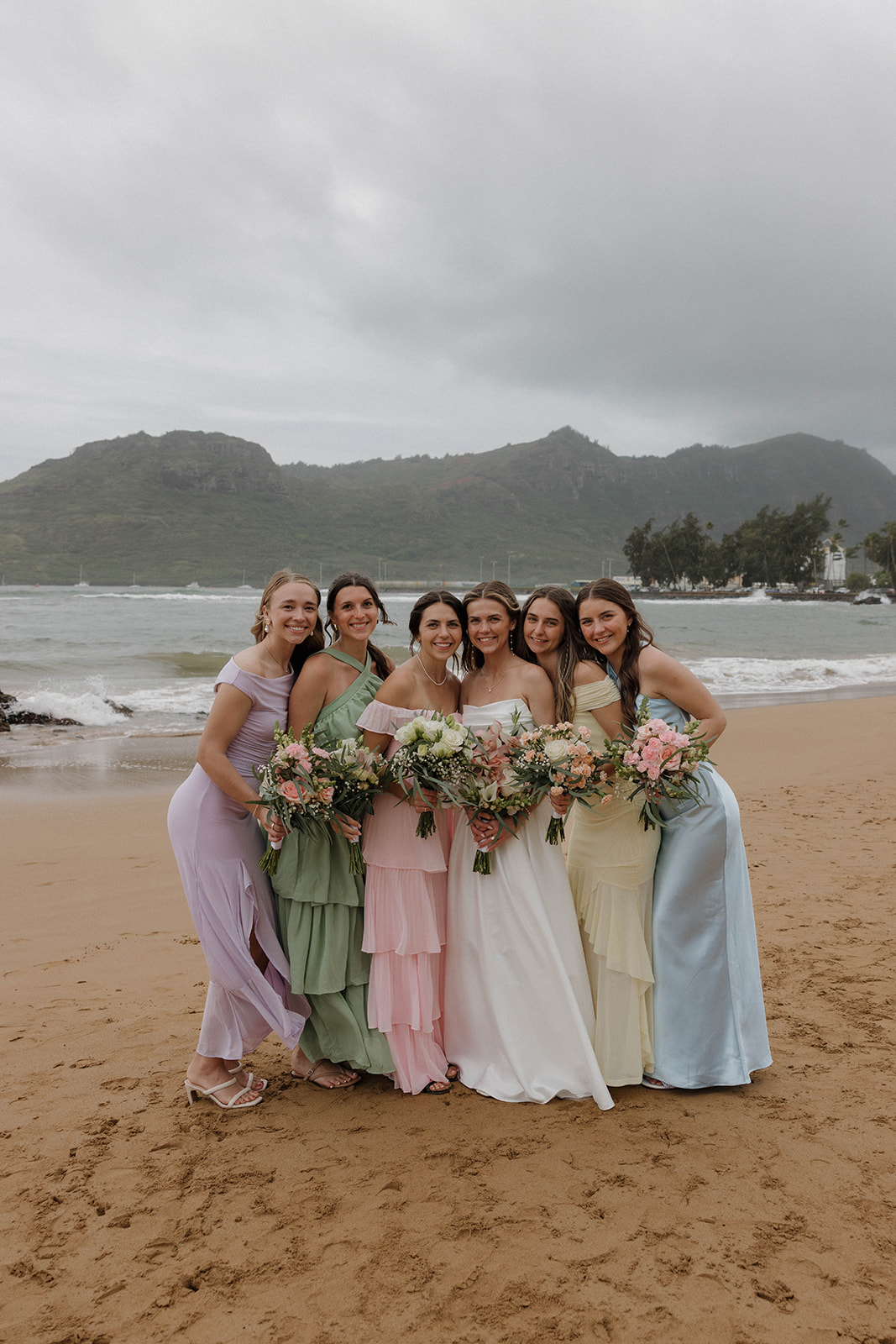 Bride and her bridesmaids in pastel dresses standing on the beach and smiling for wedding photos during a destination wedding in hawaii.