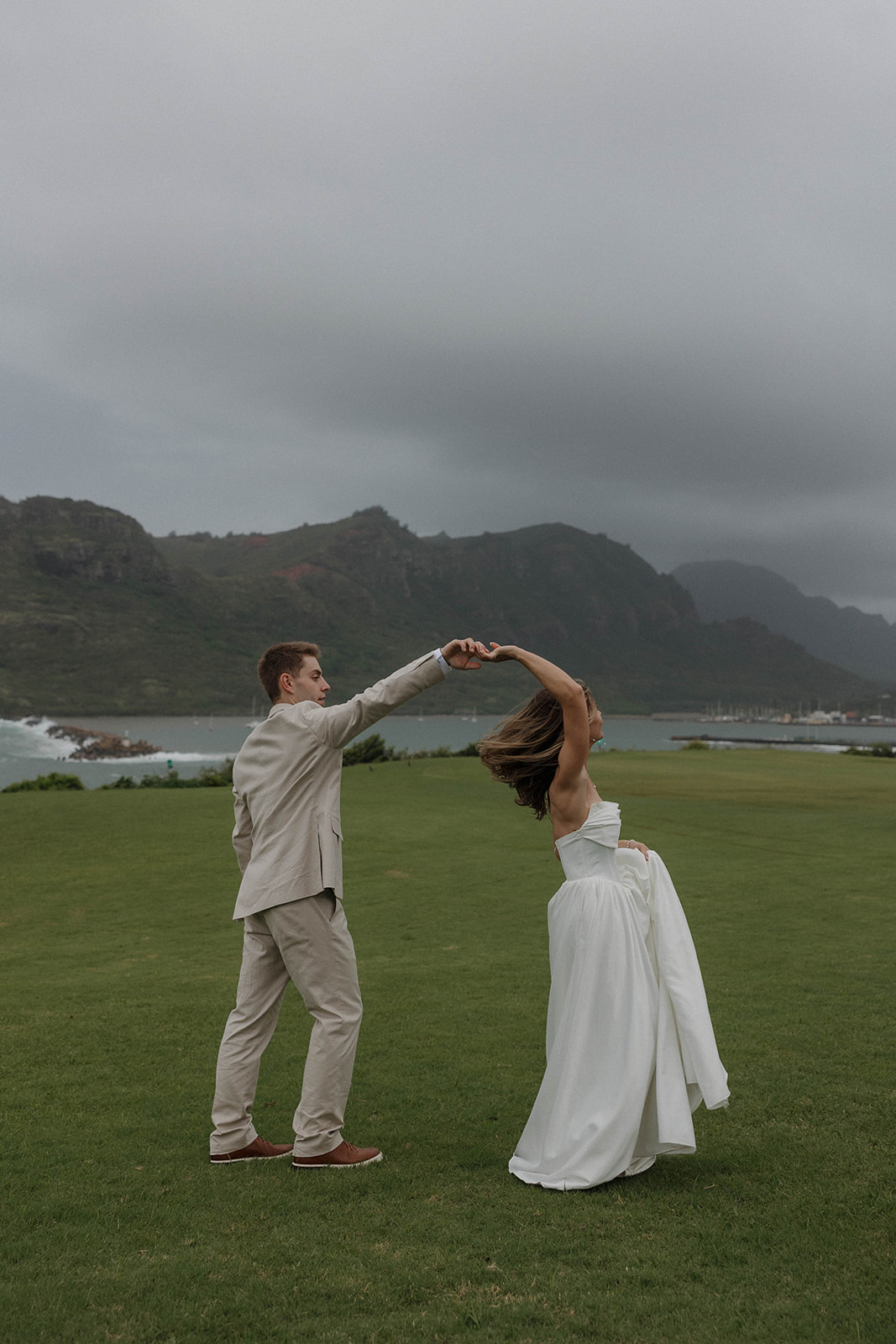 Bride and groom spinning together on a grassy cliffside overlooking the coastline at a destination wedding in hawaii.