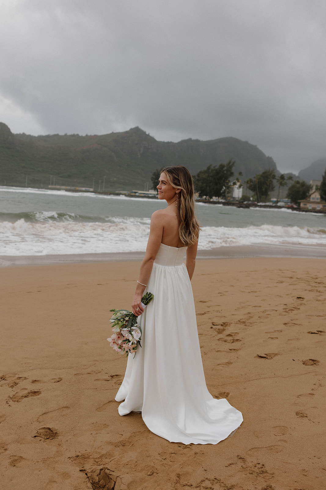 Bride standing barefoot on a sandy beach holding her bouquet with misty mountains and waves in the background.