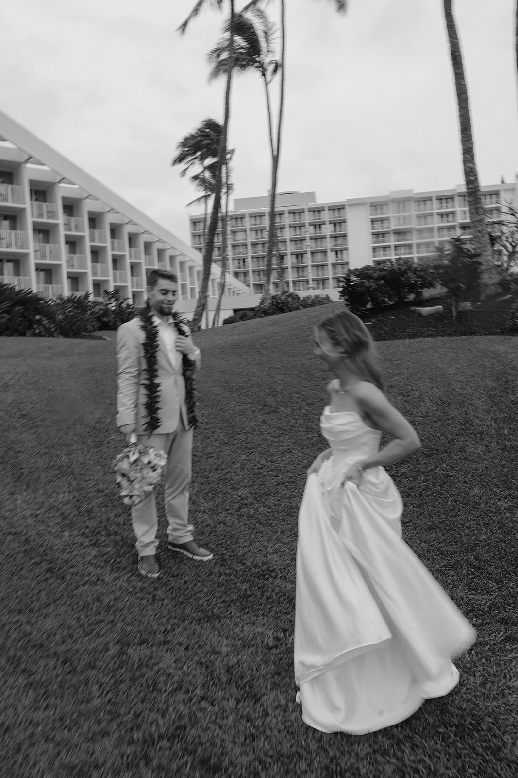 Black and white candid portrait of the bride twirling her dress while the groom watches on a tropical lawn.