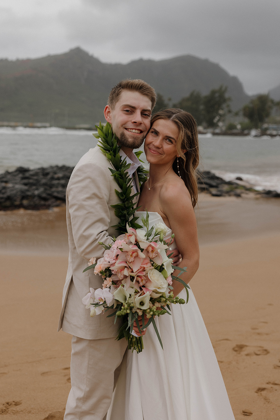 Joyful newlywed portraits on the sand with tropical mountains and stormy skies at a destination wedding in hawaii.