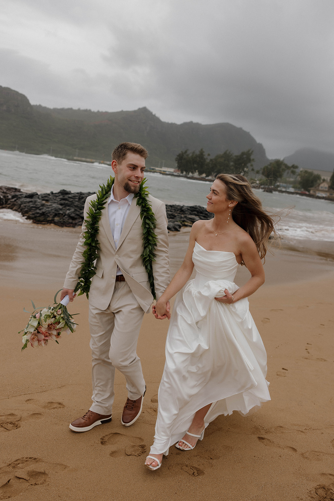 Bride and groom walking hand in hand along the beach with waves crashing behind them.