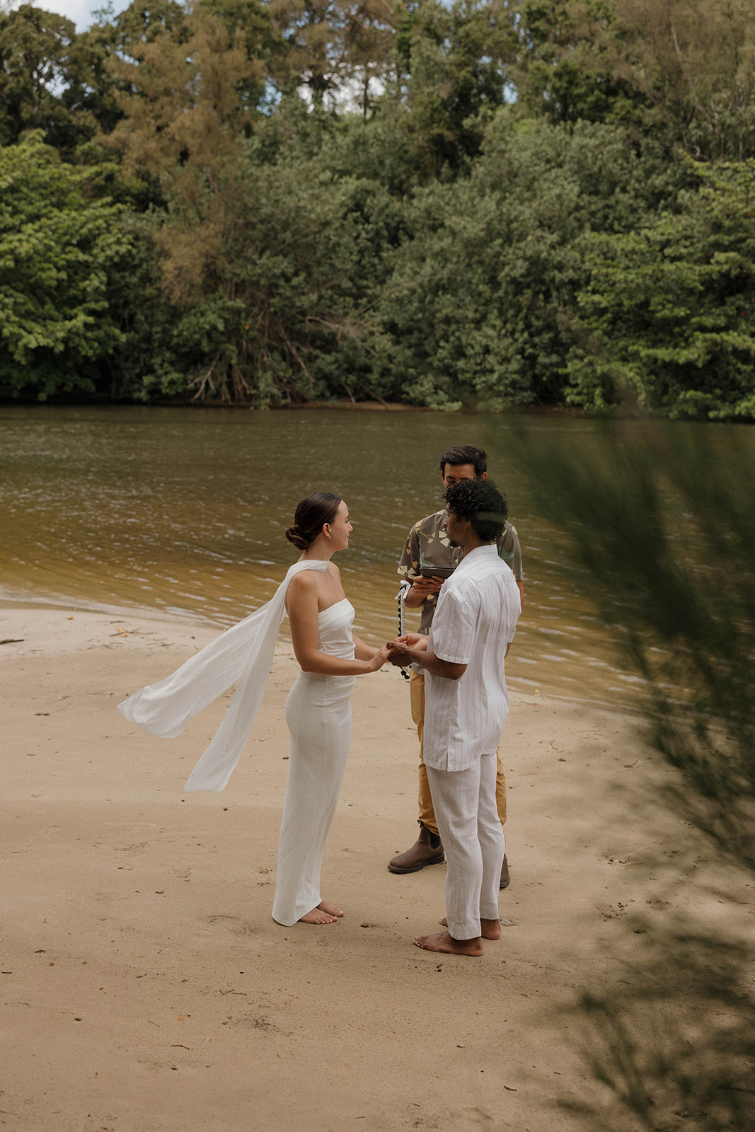 Newlyweds standing on the bank of a river in Kauai holding hands and saying their vows with the officiant standing next to them.