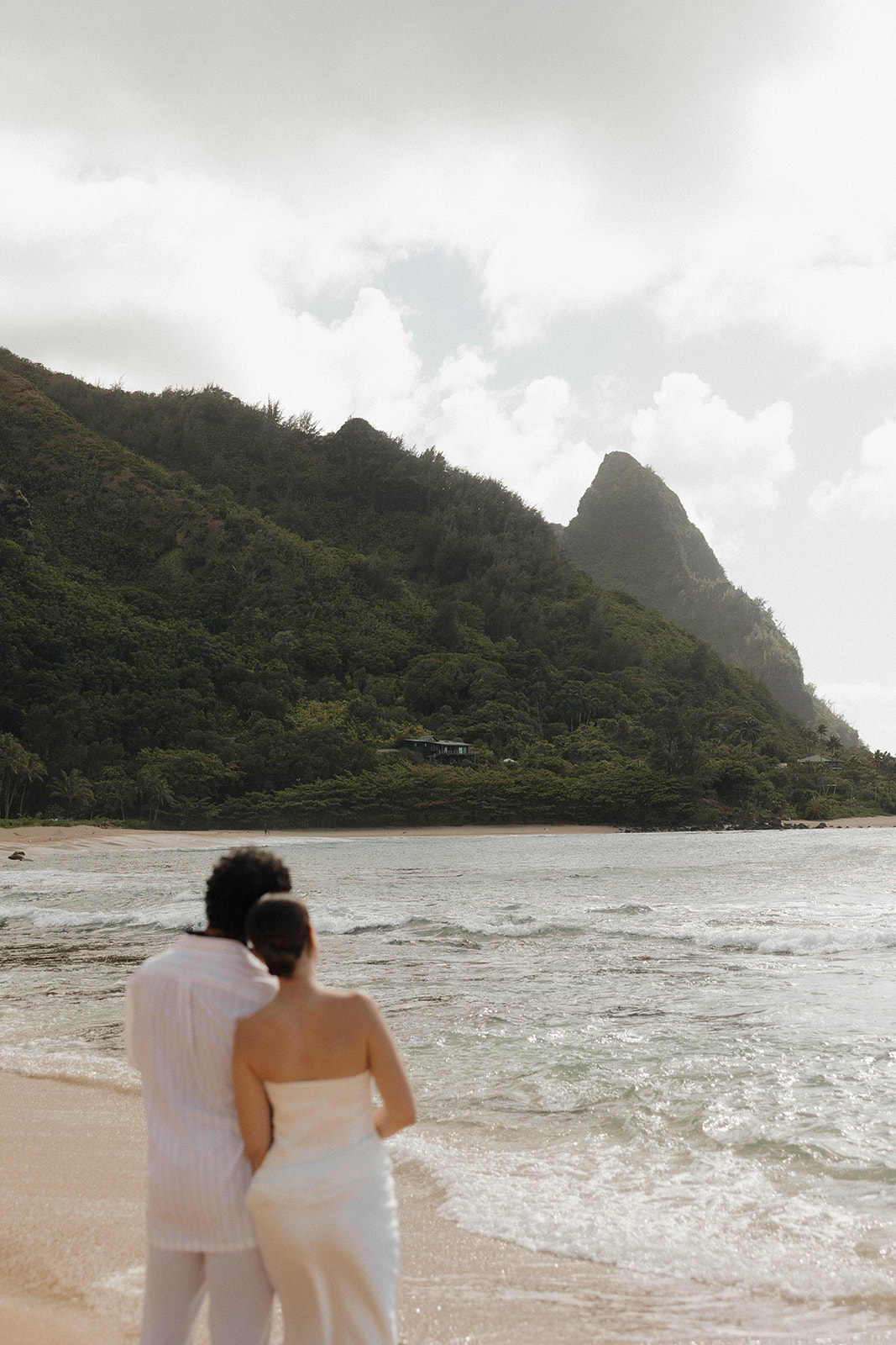 A detail photo of the bride and groom hugging into each other and standing on the beach overlooking the waves, with the background in focus and the couple blurred out.