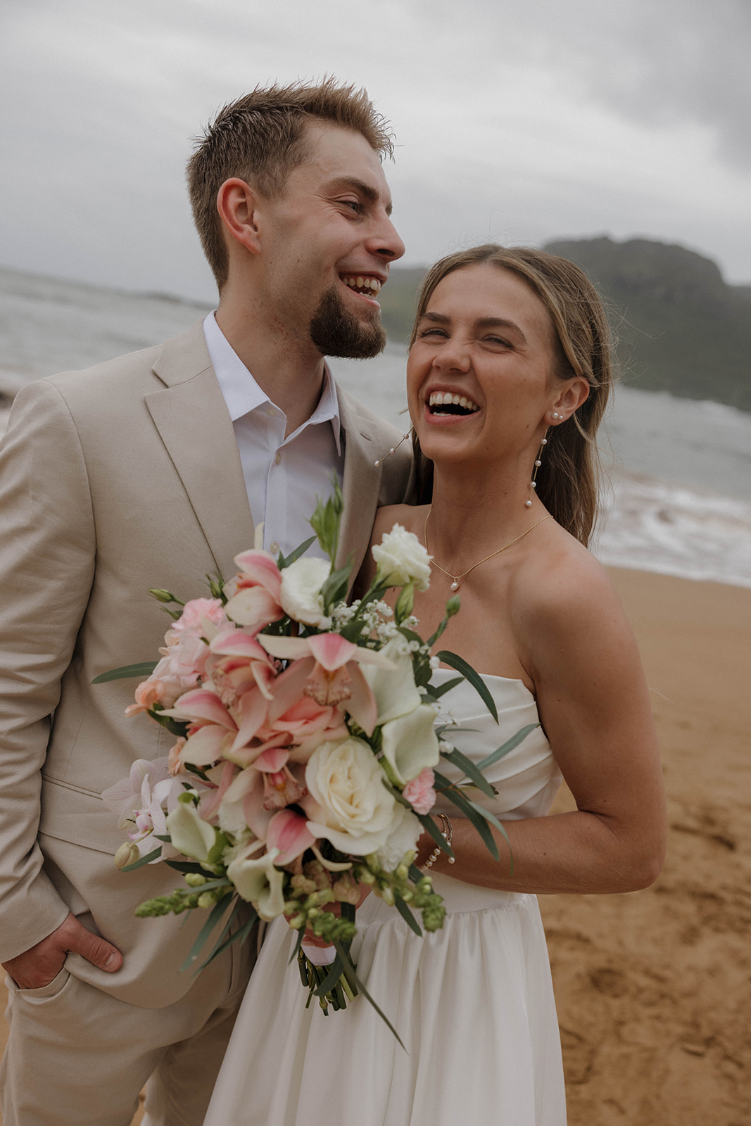 Joyful newlywed portraits on the sand with tropical mountains and stormy skies at their micro wedding in Kauai