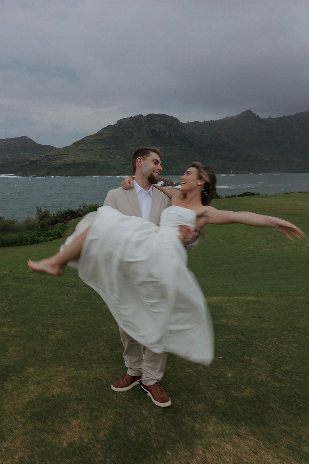 Playful newlywed portrait of the groom spinning the bride in his arms on a coastal lawn.