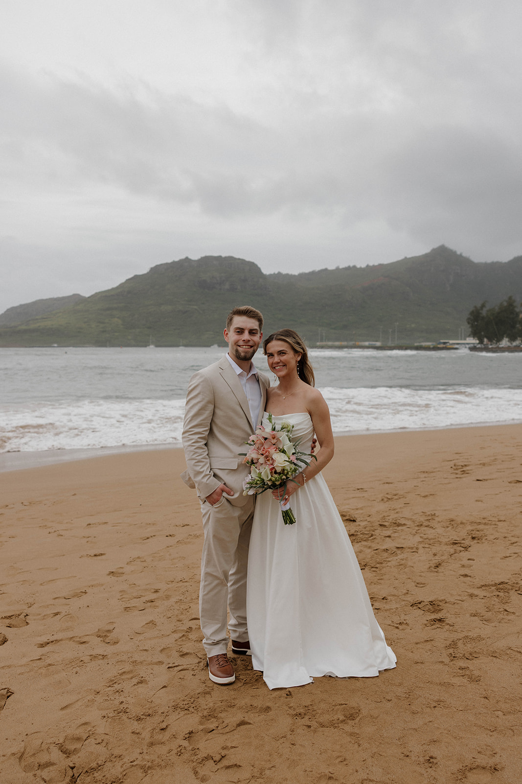 Joyful newlywed portraits on the sand with tropical mountains and stormy skies at their micro wedding in Kauai