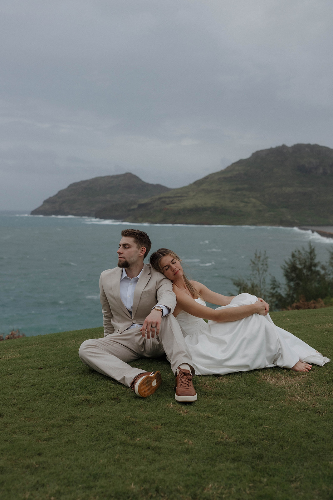 Bride and groom sitting together on a grassy bluff with waves crashing below them.