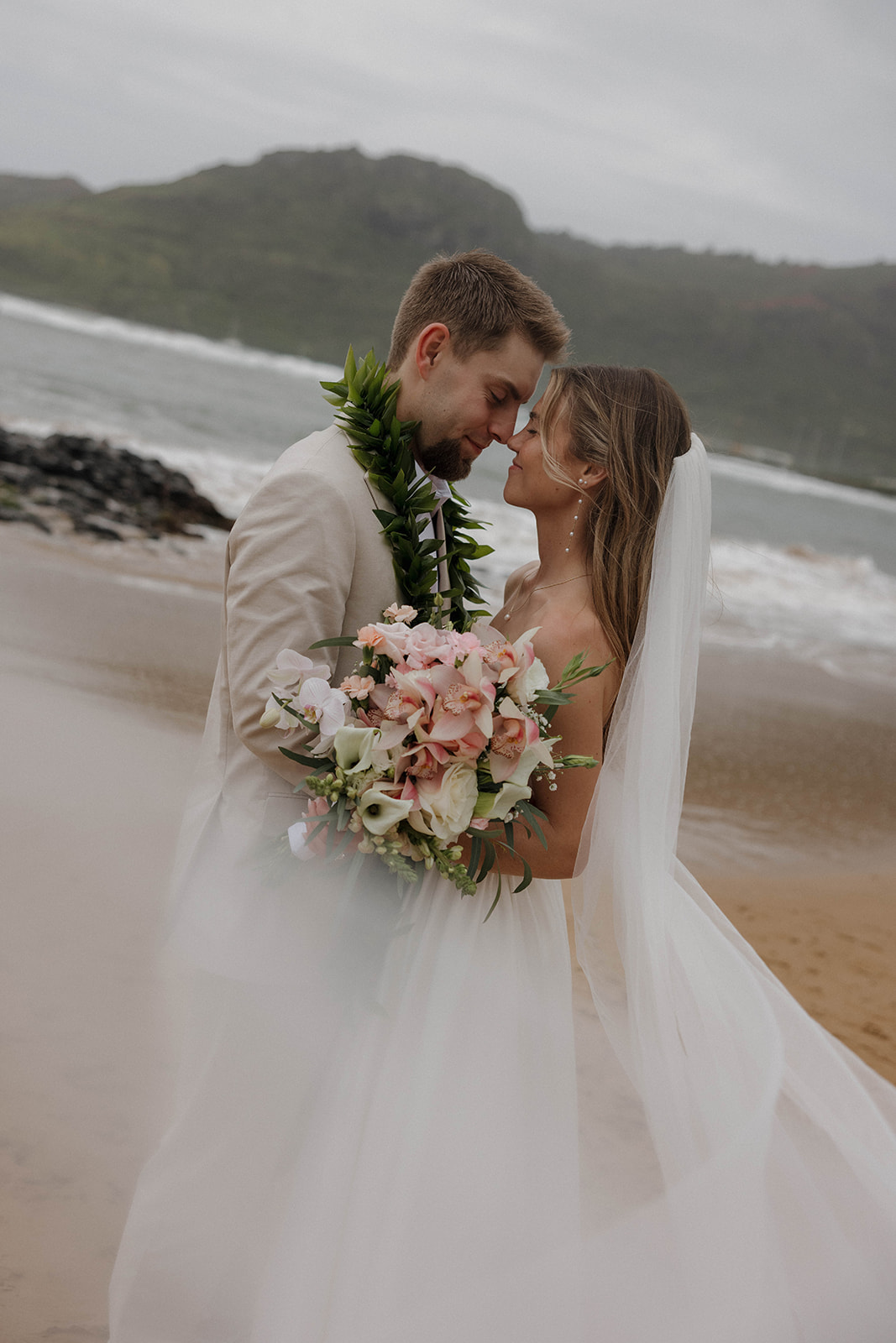 Outdoor beach portraits of the bride and groom embracing with the brides veil flowing in the foreground with misty mountains behind them during their Micro Wedding in Kauai