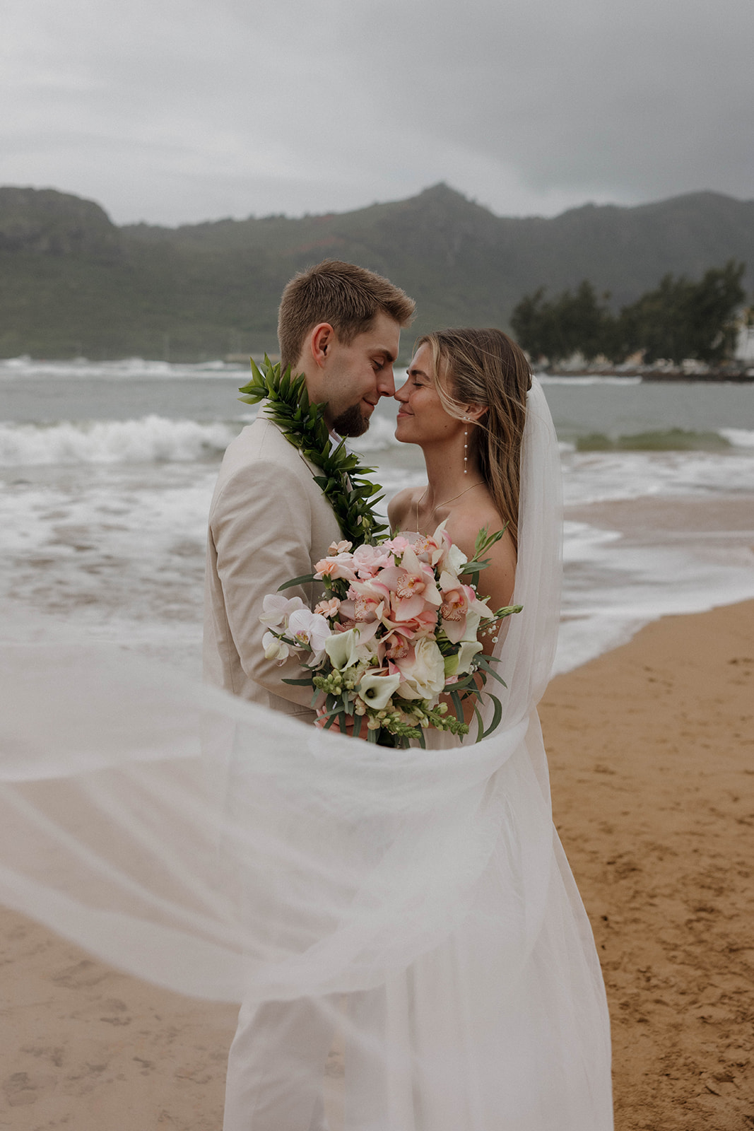 Romantic veil-wrapped portrait of the bride and groom embracing on a windy beach.