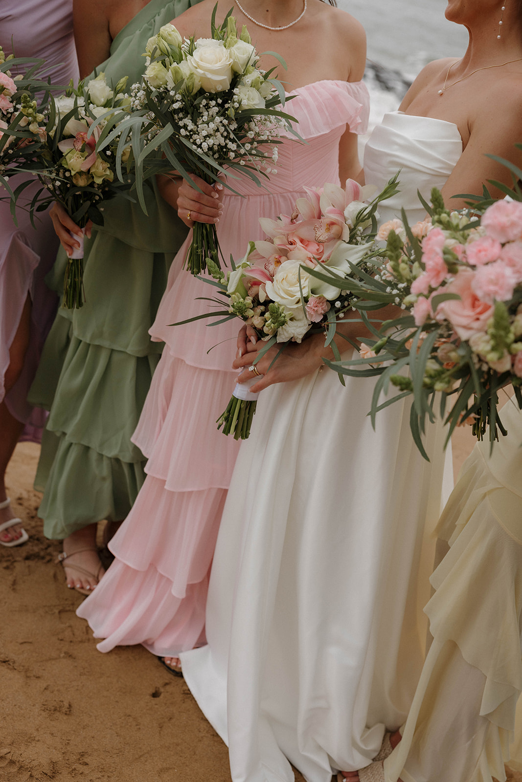 A detail photo of the bride and bridesmaids dresses, with the, all holding colorful wedding bouquets on the beach.