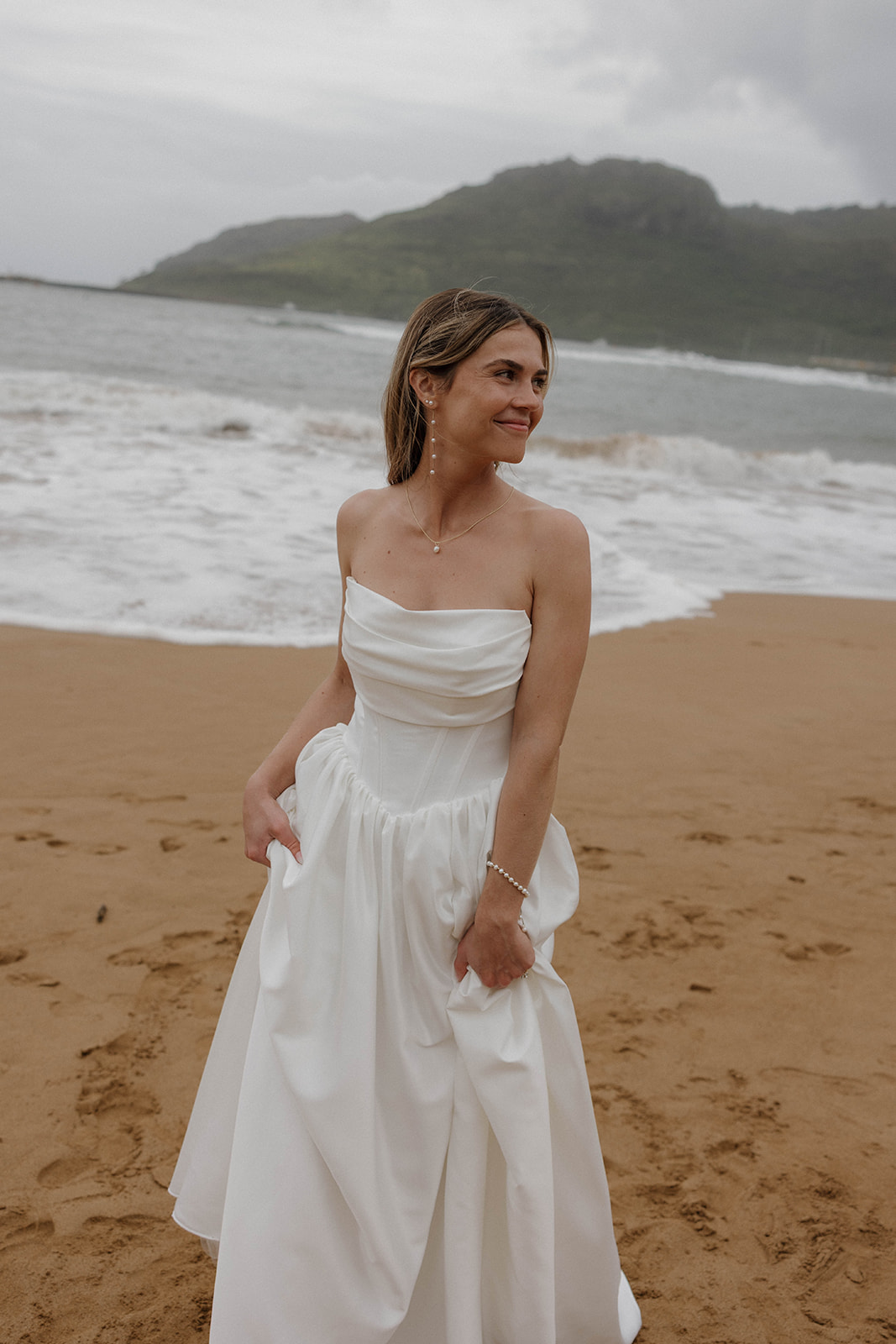 Bride standing barefoot on a sandy beach holding her dress with misty mountains and waves in the background.