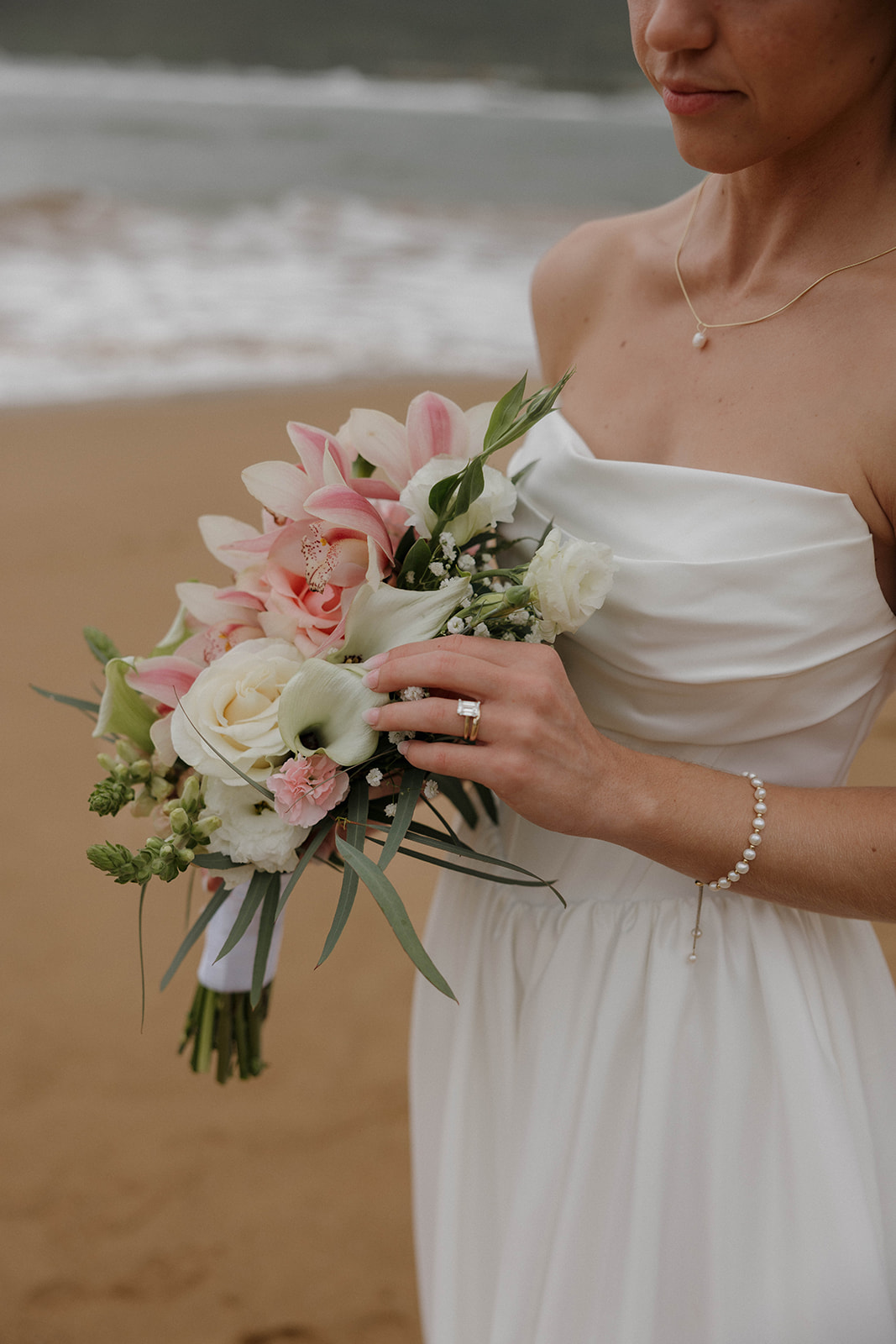 Bride standing barefoot on a sandy beach holding her bouquet with misty mountains and waves in the background.