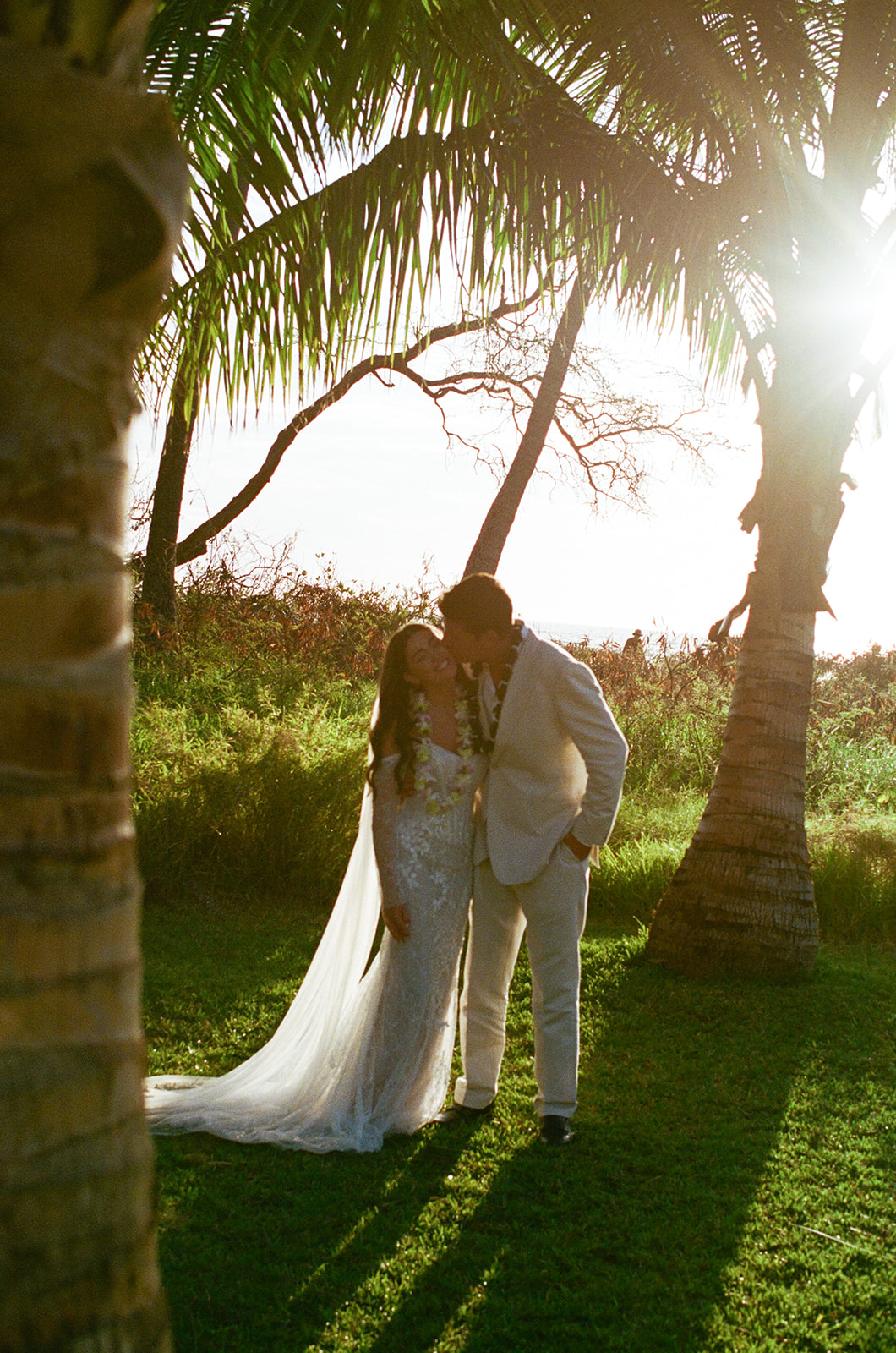 Bride and groom kissing under palm trees backlit by the setting sun in Hawaii
