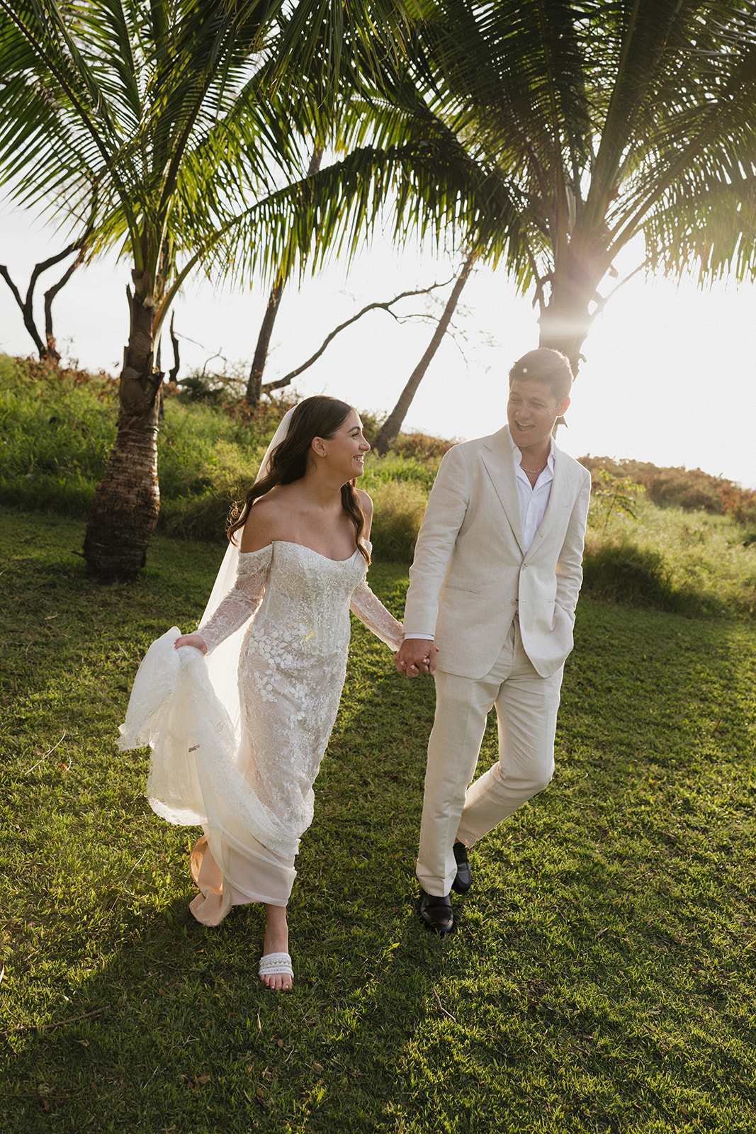 Bride and groom laughing and walking hand in hand through a sunlit Hawaii lawn after their wedding
