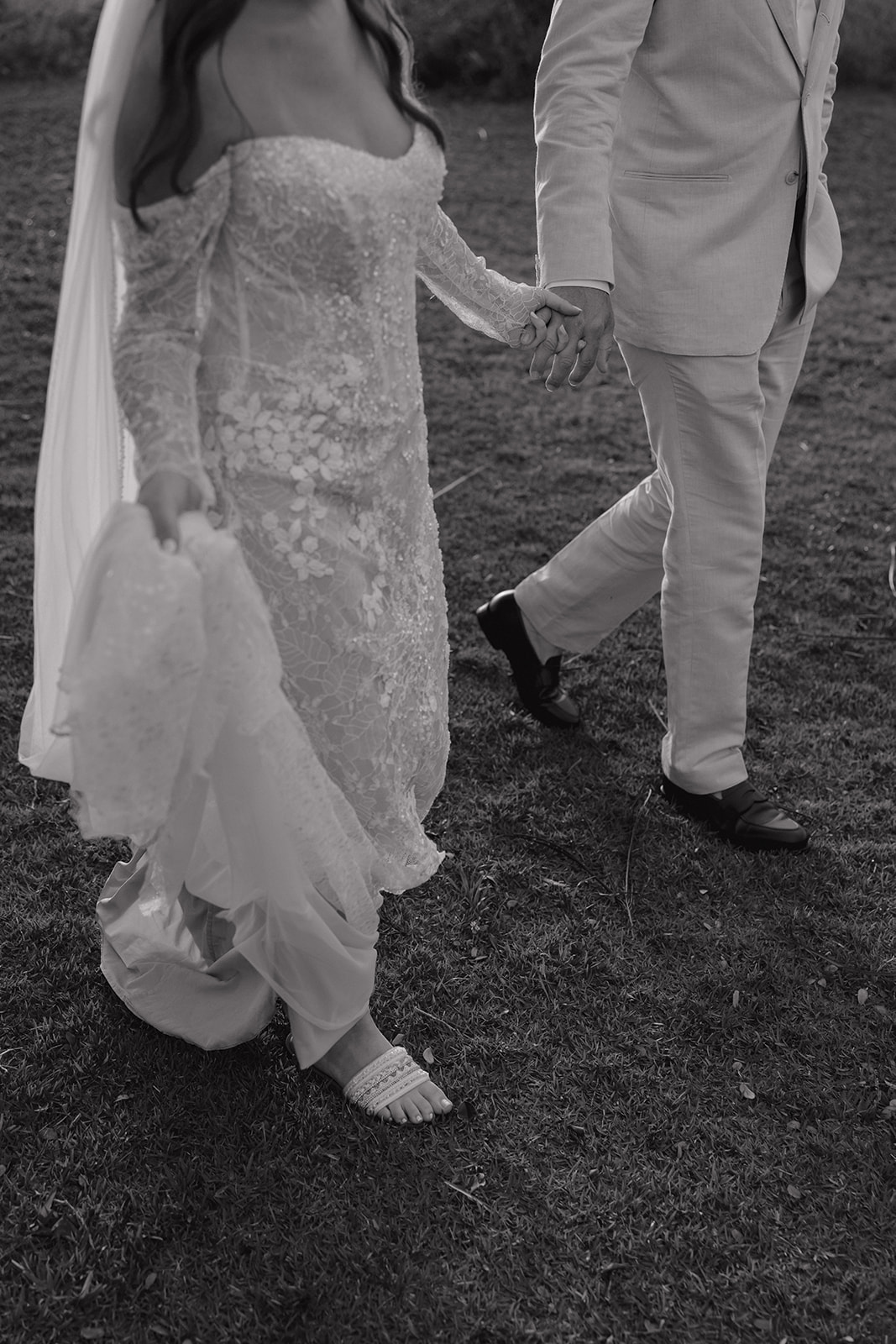 Black and white close-up of bride in lace gown and groom holding hands while walking across the grass
