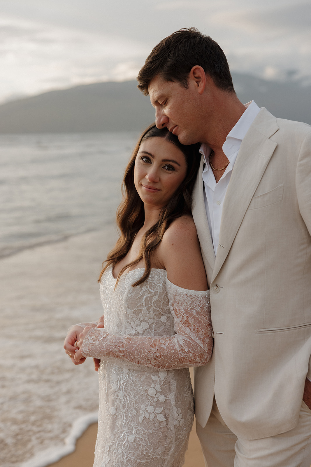 Bride leaning against groom on Maui beach at dusk with mountain backdrop
