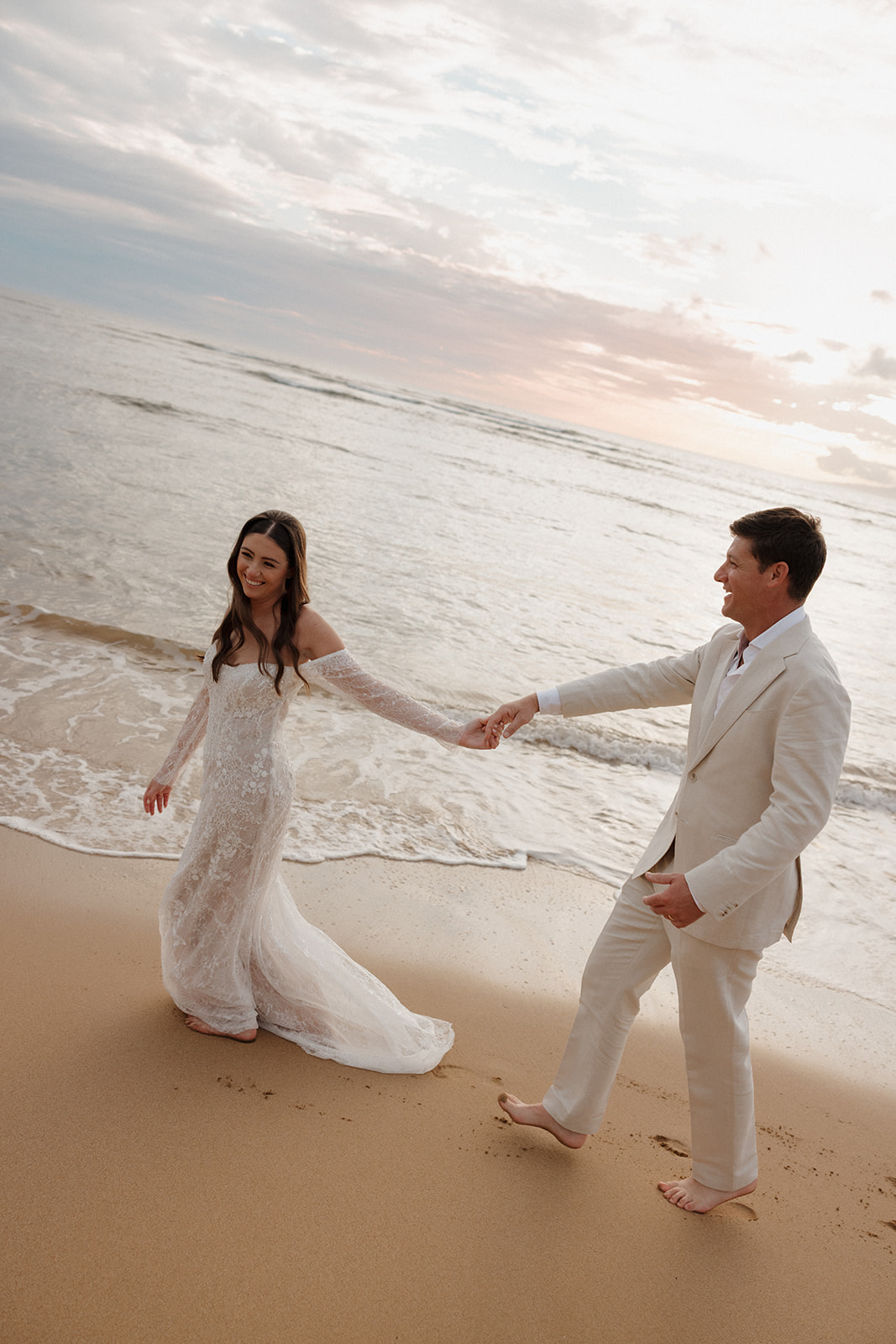 Bride and groom laughing and holding hands on the beach at sunset
