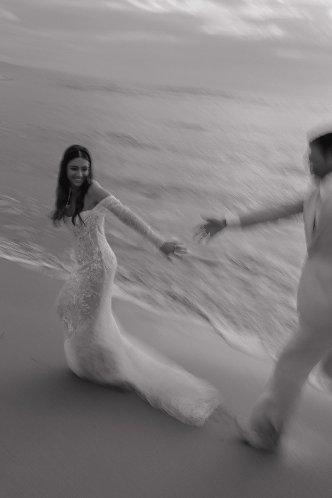 Artistic blurred black and white photo of bride laughing while reaching for groom on the beach
