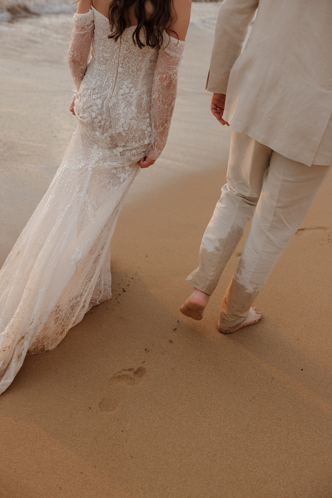 Close-up of bride's lace wedding dress train and groom's sandy bare feet walking on the beach
