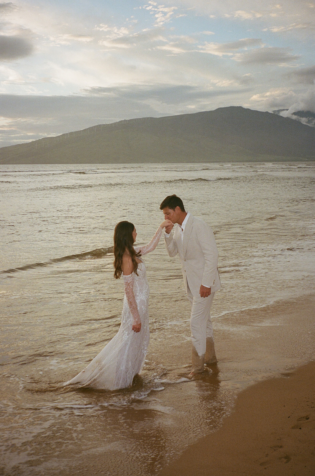 Groom kissing bride's hand while standing in the surf during their Maui elopement at sunset
