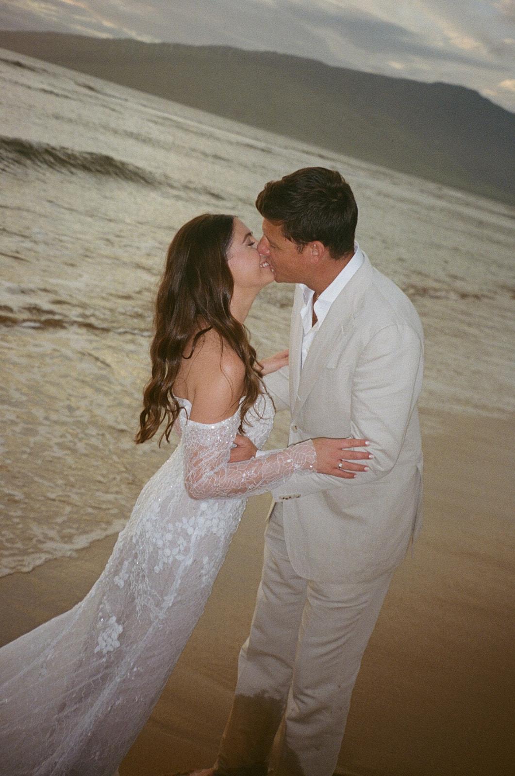 Bride and groom sharing a kiss on the beach at dusk with ocean waves behind them
