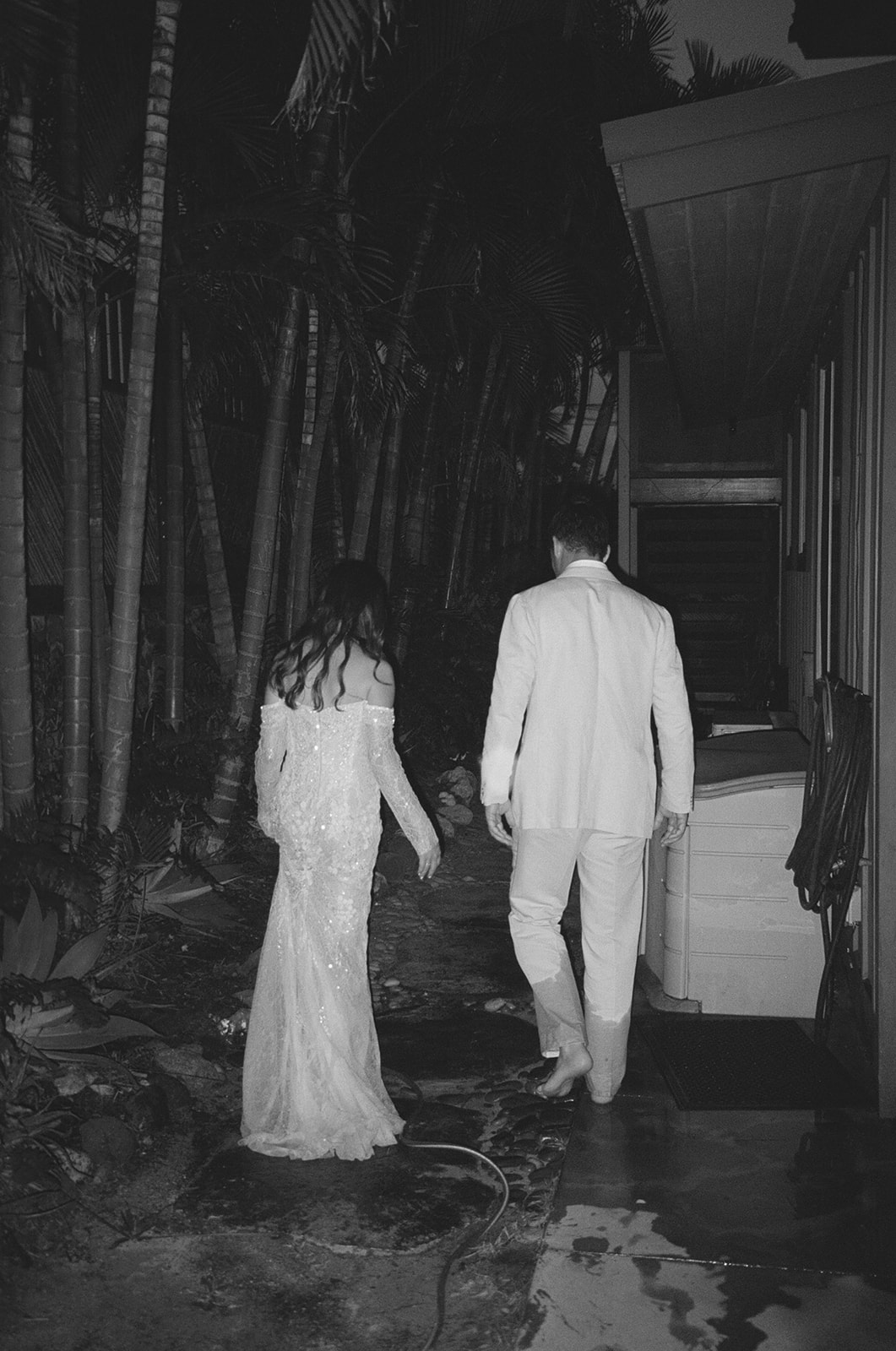 Black and white photo of bride and groom walking together at night through tropical Hawaii grounds

